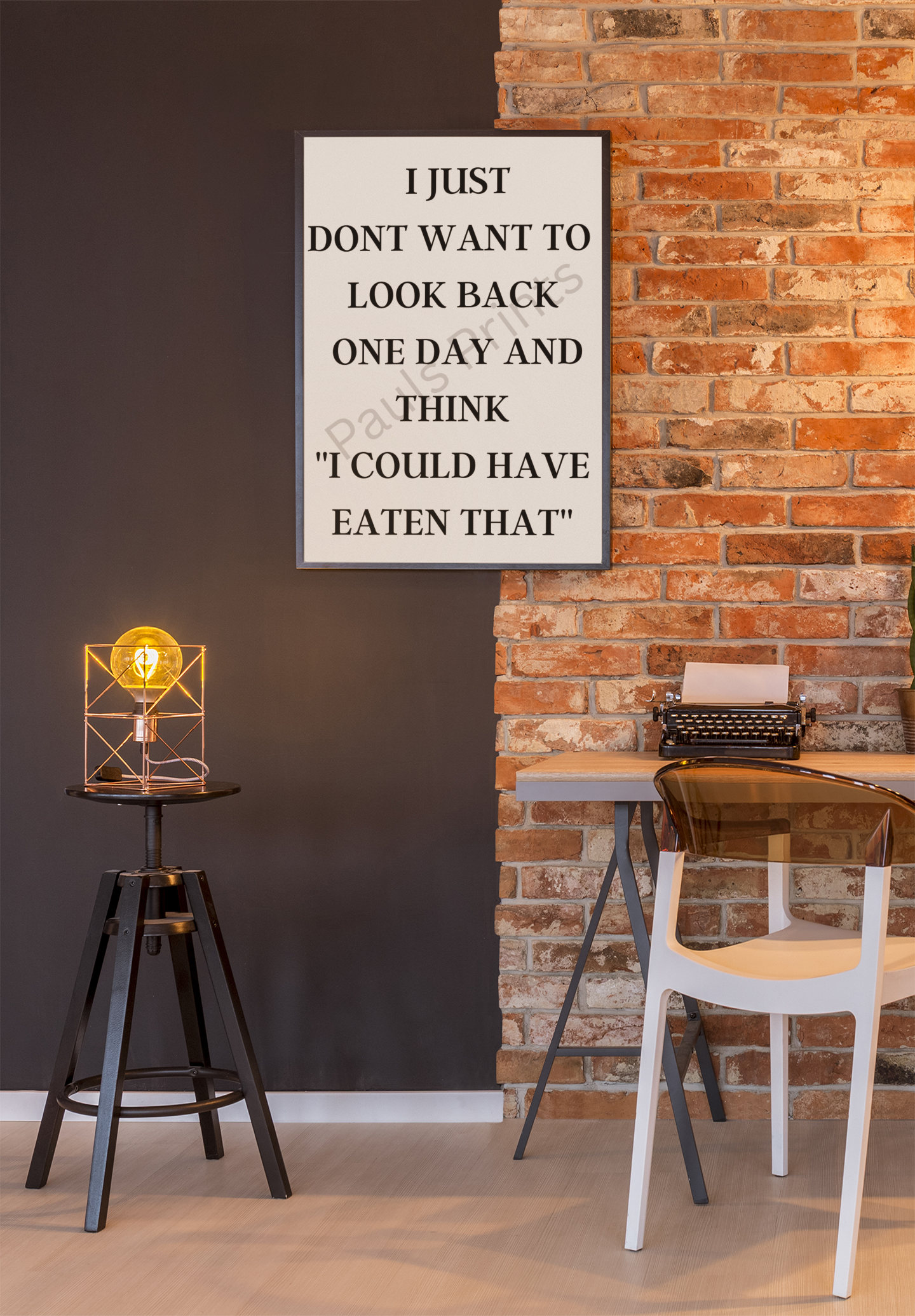 Framed print with the quote “I just don’t want to look back one day and think I could have eaten that” on a brick and dark wall in an industrial-style workspace with a vintage lamp.