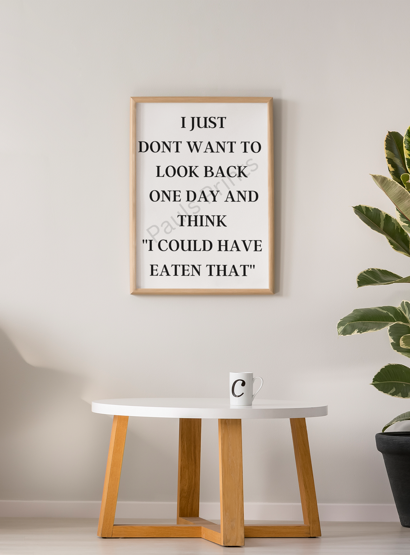 Wooden-framed print saying “I just don’t want to look back one day and think I could have eaten that” on a neutral wall above a small white table and coffee cup.