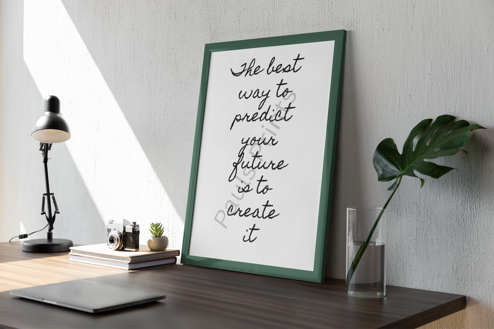 Motivational quote print in green frame leaning against a textured wall beside a desk with modern lamp, camera, and leaf in a vase.