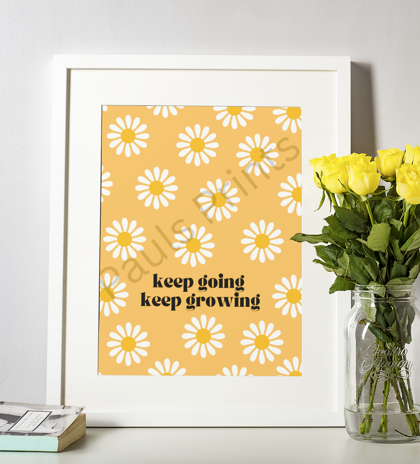 Framed wall art with white daisies on a yellow background and the motivational phrase "keep going keep growing", displayed next to a bouquet of yellow roses in a glass jar.