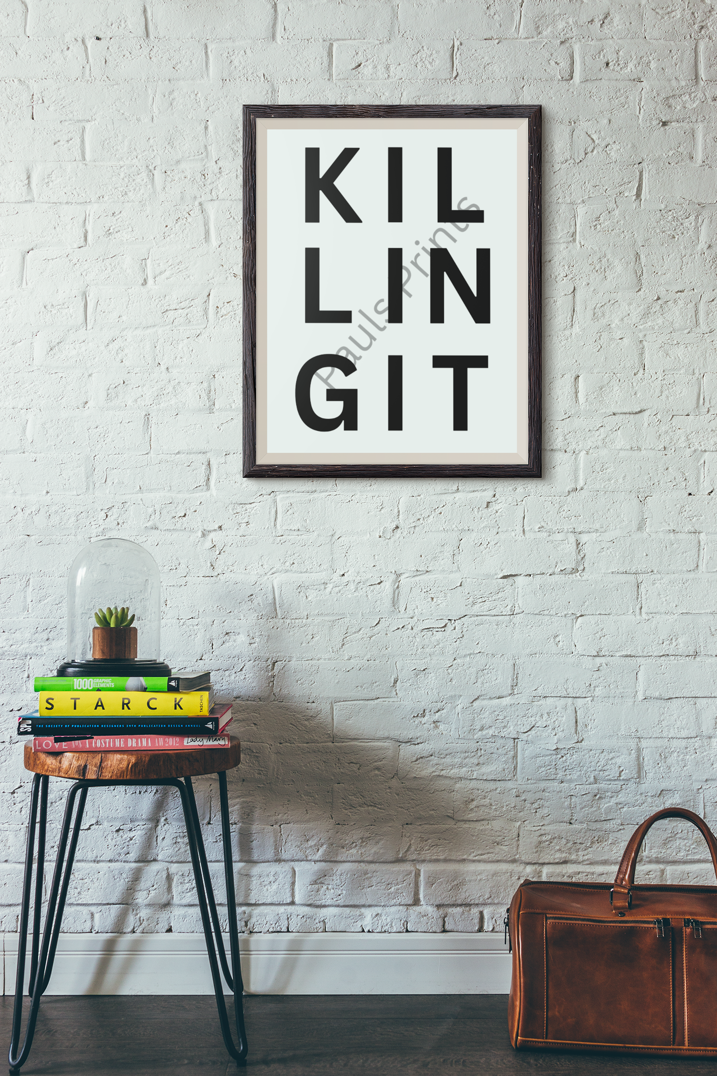 Motivational “Killing It” print displayed on a white brick wall above a stack of colourful books and a leather bag in a creative home workspace.