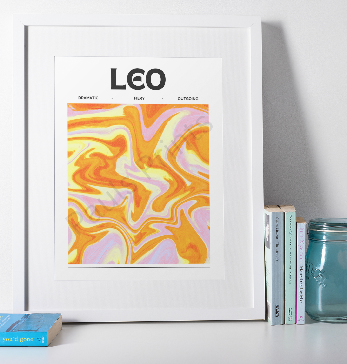 Colorful Leo horoscope print framed in white, showcasing energetic abstract swirls and placed next to a small stack of books and a teal jar.
