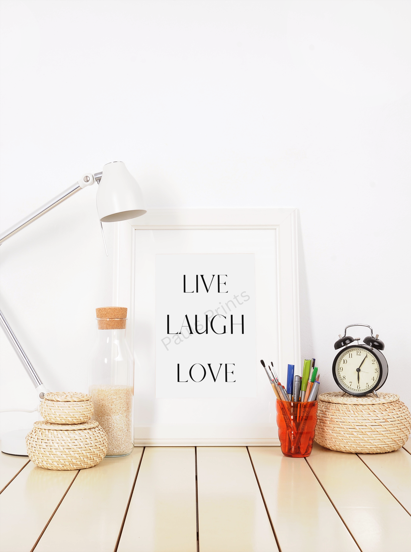 White-framed Live Laugh Love print on a desk with lamp, woven baskets, glass bottle, clock, and pens in a bright workspace.