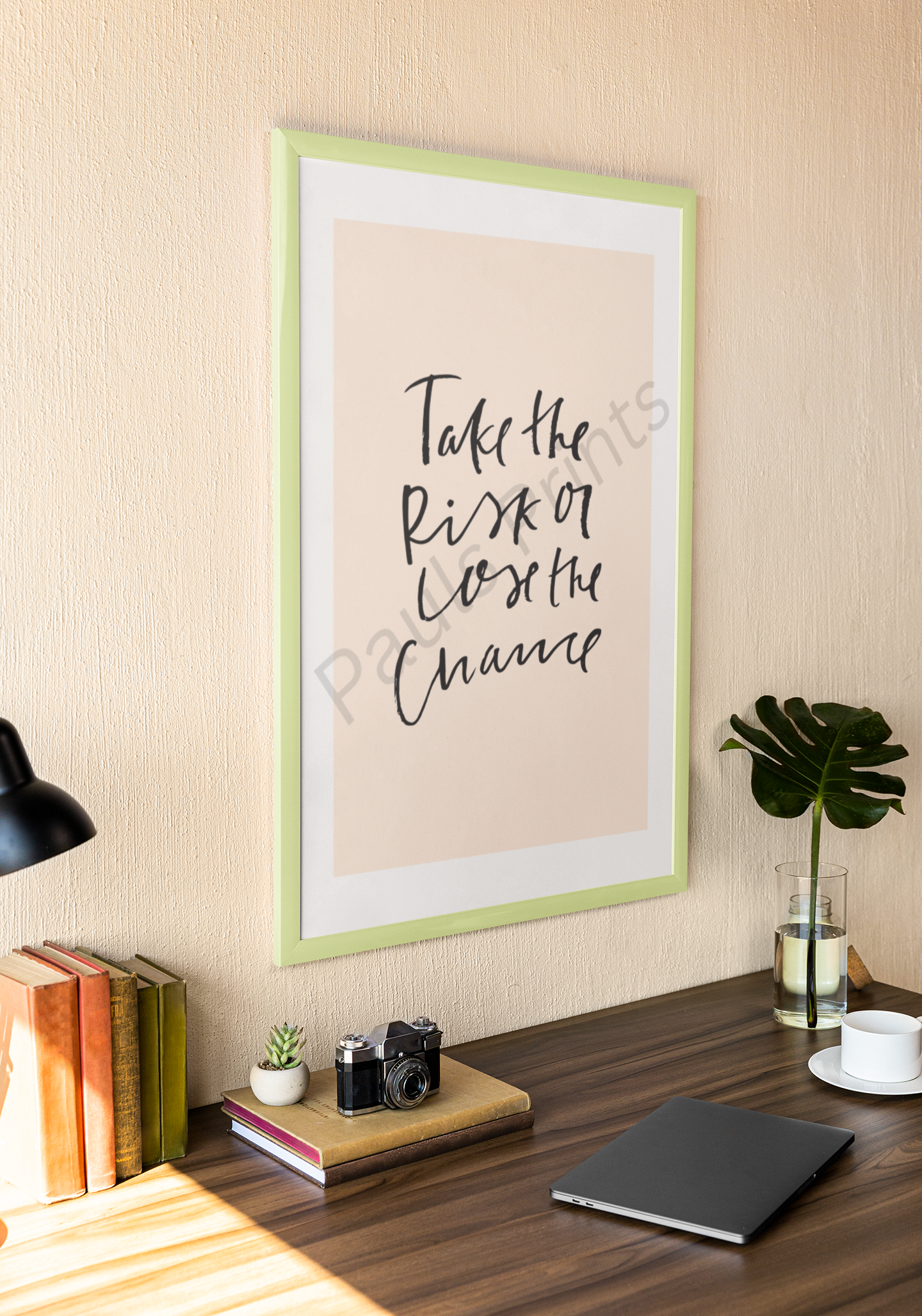 Cozy desk space with framed motivational print reading “Take the risk or lose the chance” in black handwritten script on beige background, decorated with books, a plant, and a camera.