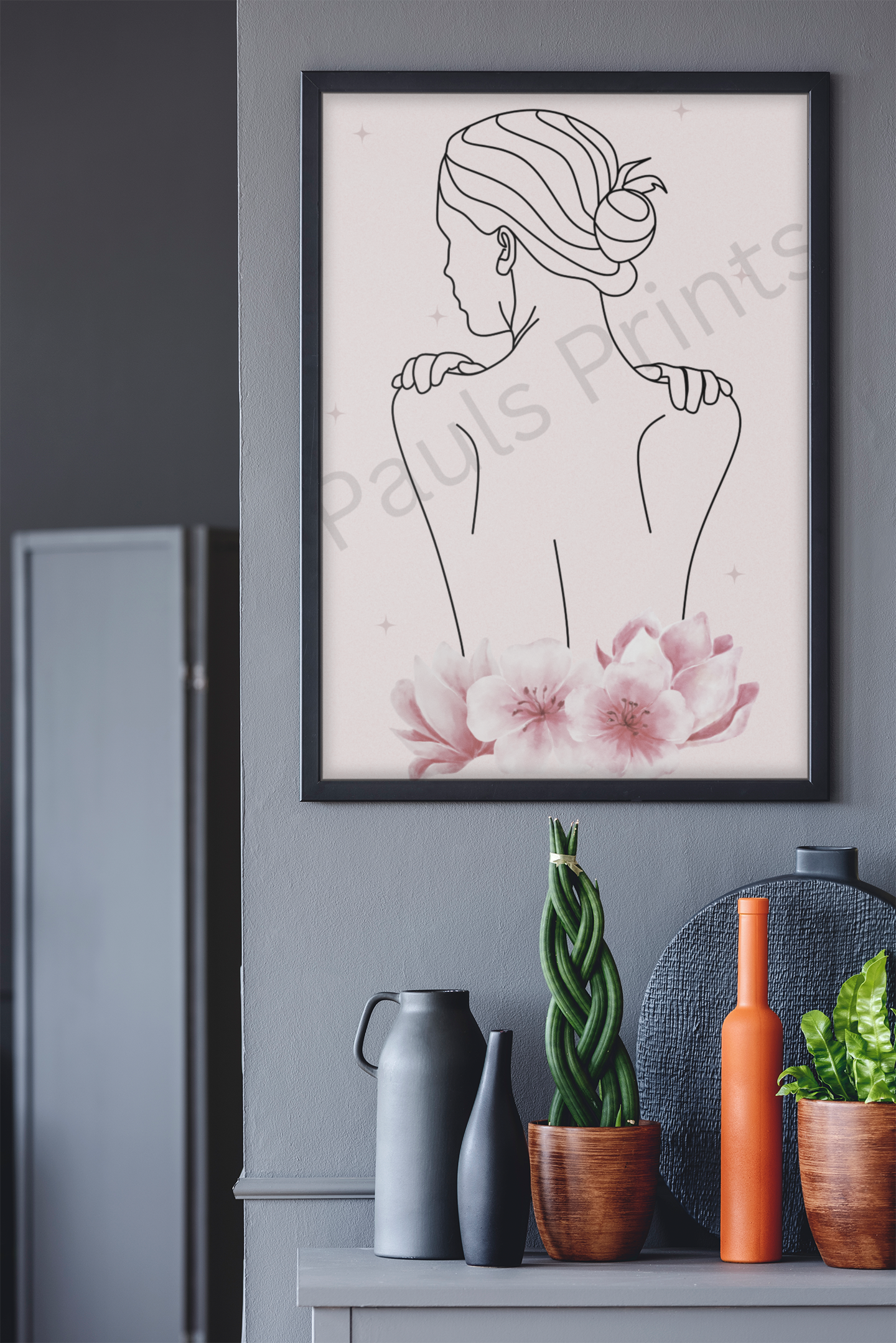 Black-framed minimalist line art print of a woman with a bun, paired with pink flowers, mounted on a dark grey wall above decorative vases and plants.