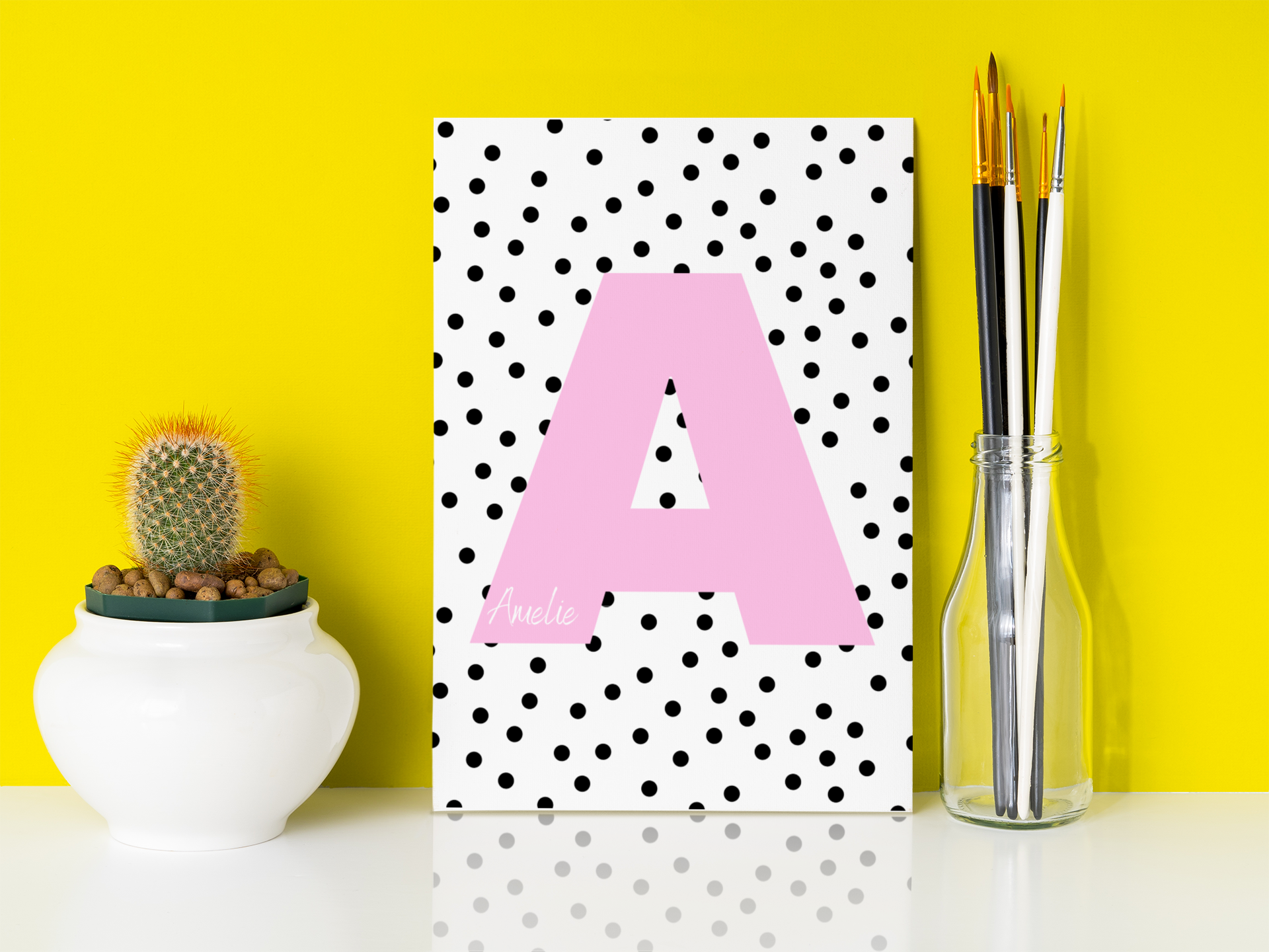 Children's initial print with pink letter A and name Amelie, set against a bright yellow background beside a cactus and paintbrush jar.