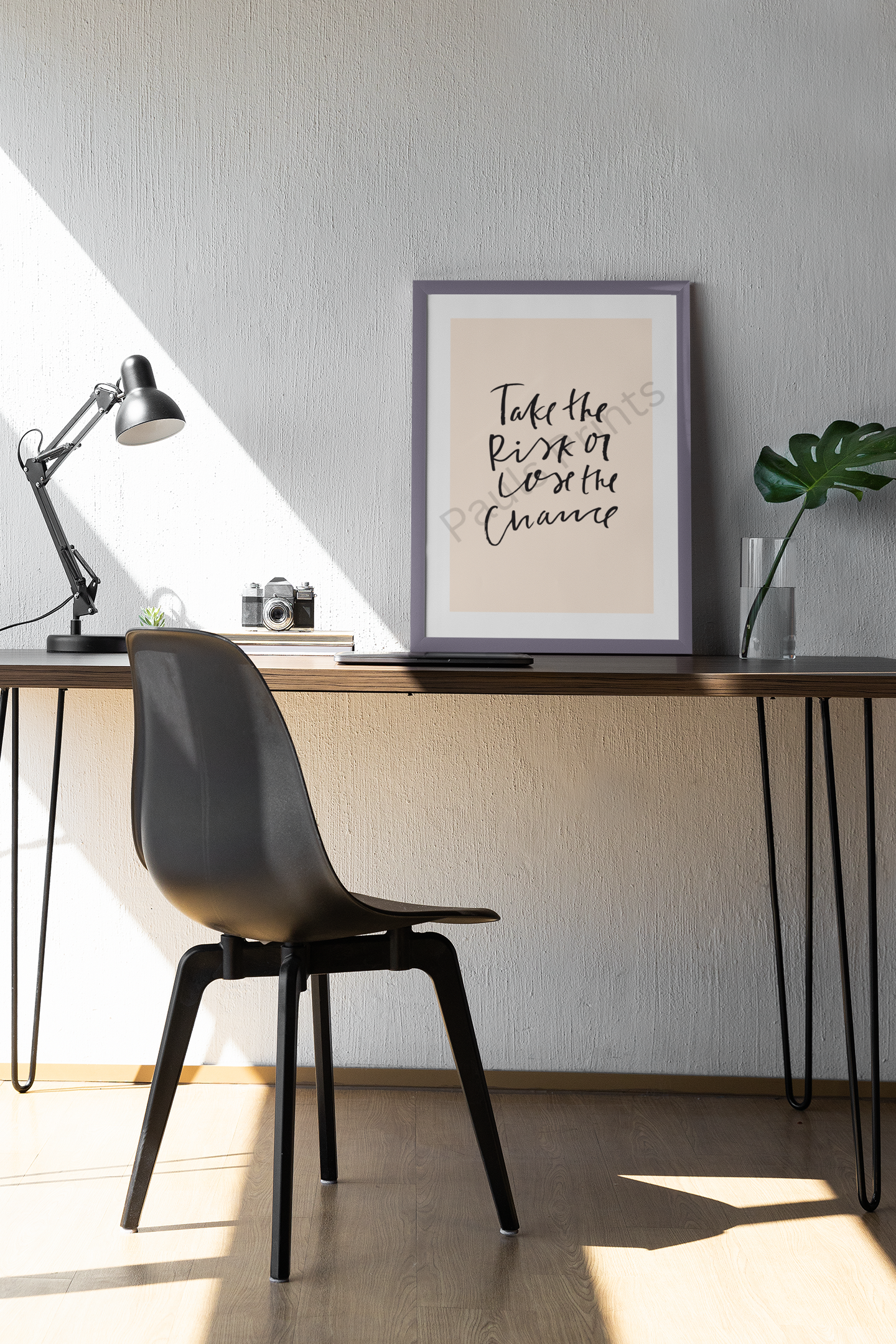Stylish modern workspace with framed motivational print reading “Take the risk or lose the chance” in black handwritten script on beige background, beside a desk lamp and green plant.