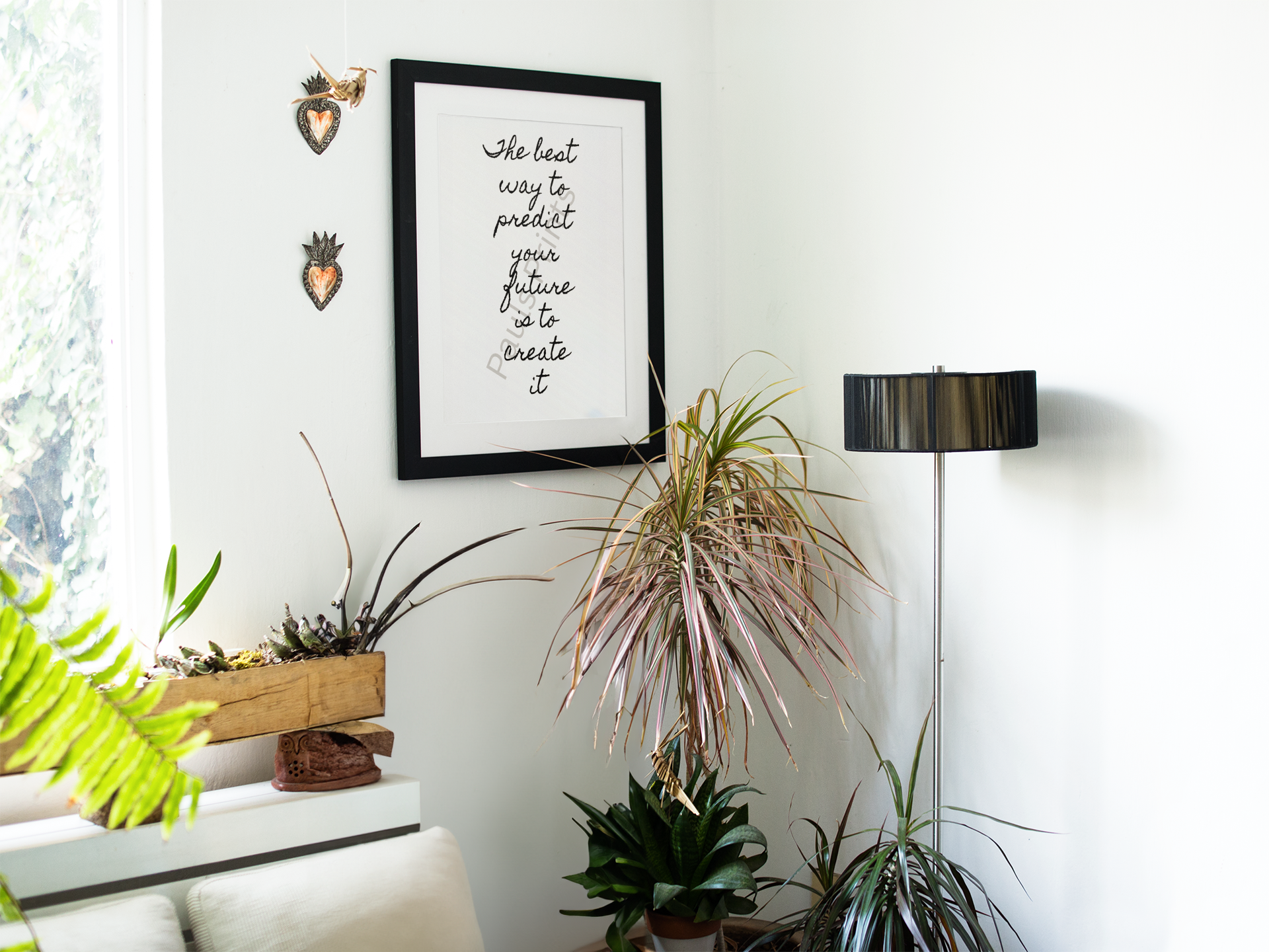 “Create your future” quote print in black frame hanging on white wall surrounded by potted plants and natural light.