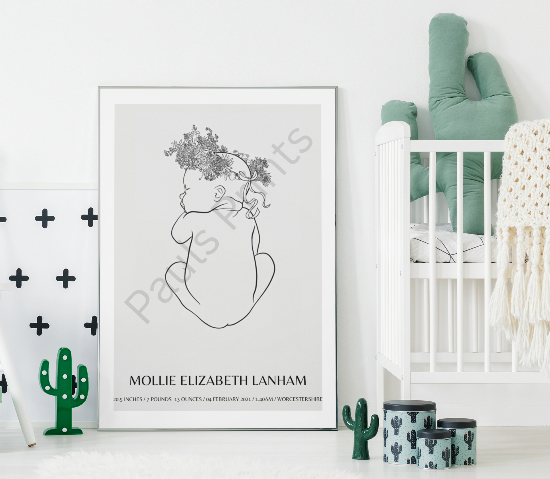 Oversized baby line art print leaning beside a cot in a neutral nursery with cactus-themed décor and white walls.