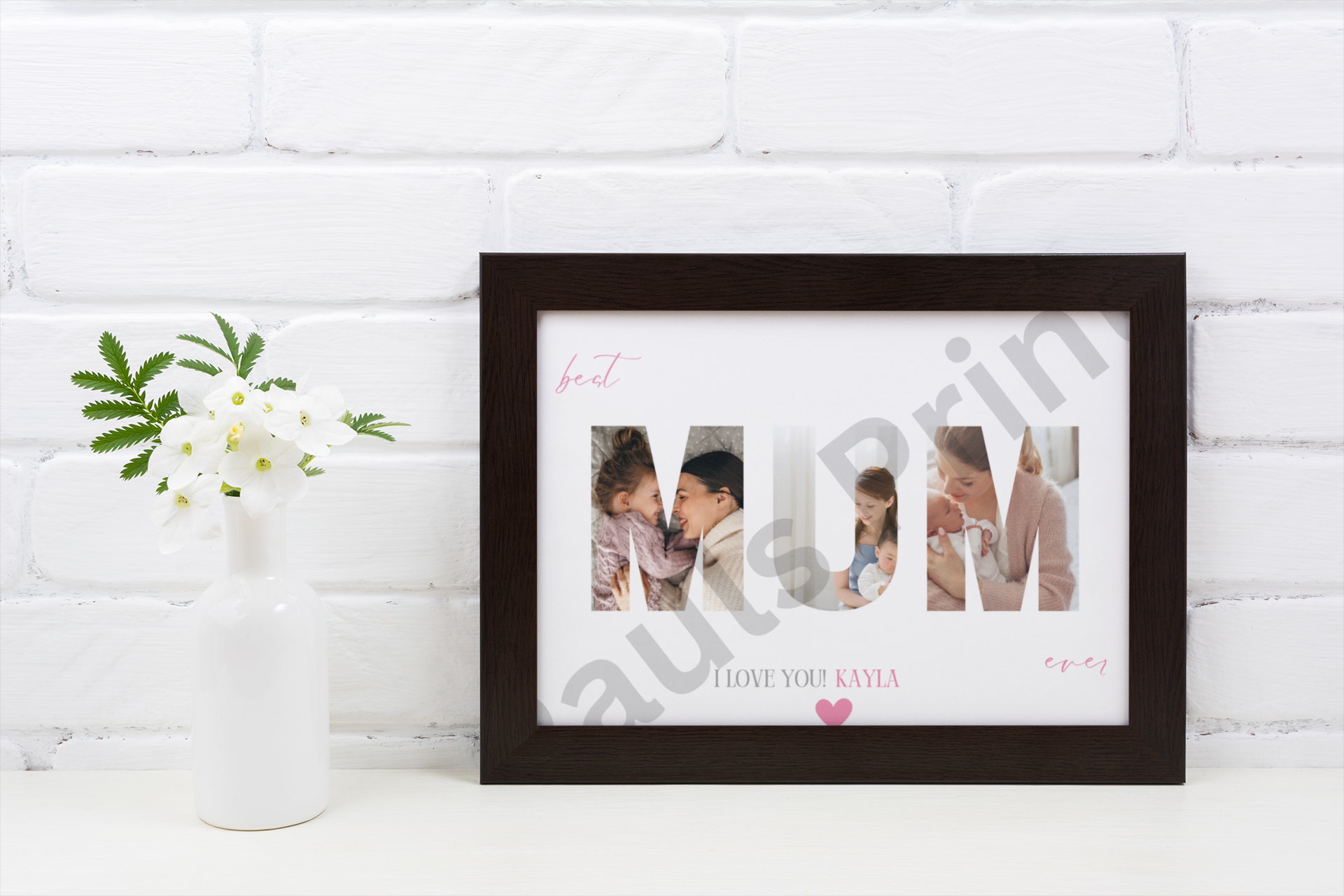 Black frame with MUM collage artwork showing family photos and personalised name, displayed on a white brick wall beside a vase of white flowers.