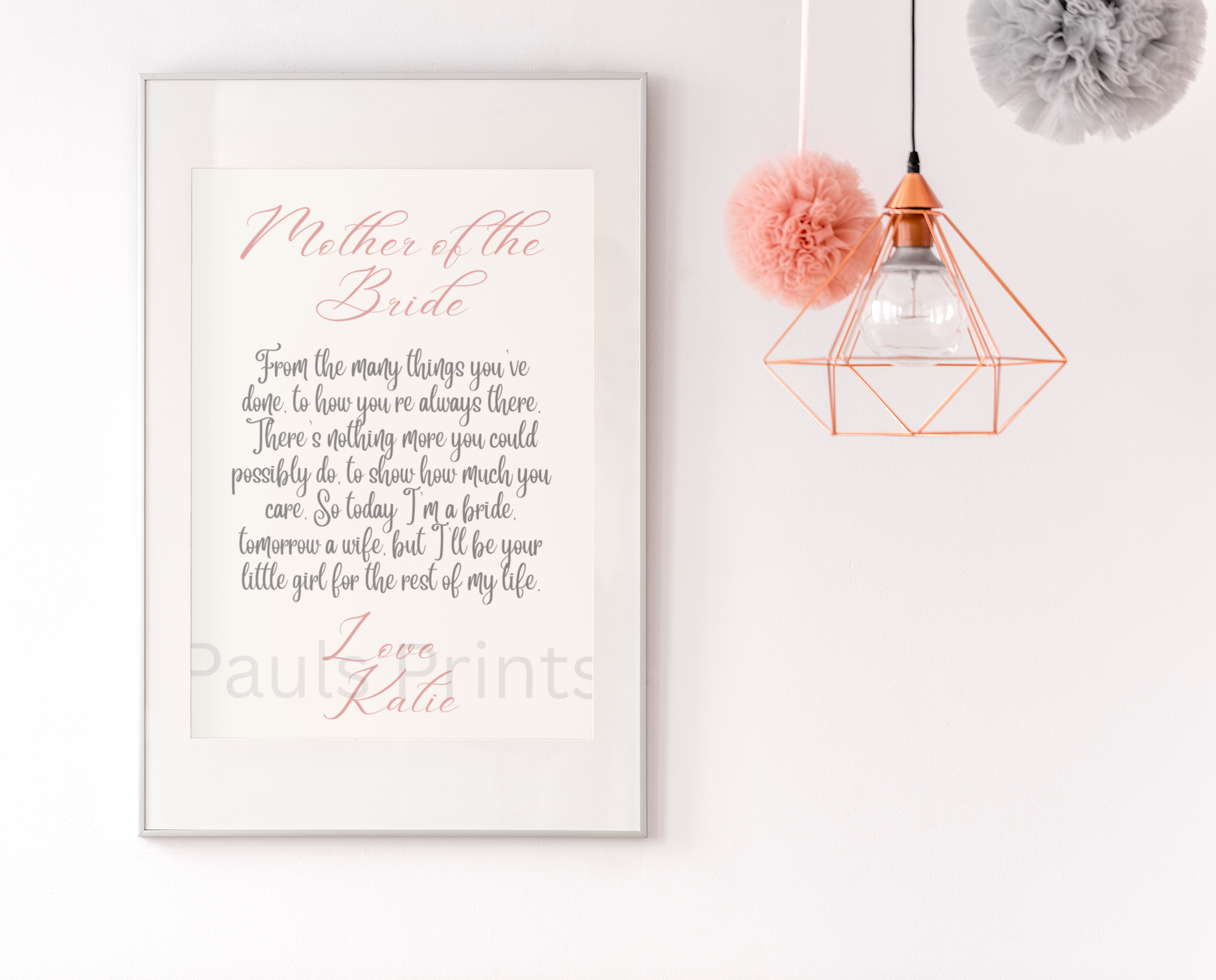 Framed wedding poem print with soft pink accents hanging on a white wall, complemented by pastel pom-pom decorations and a geometric pendant light.