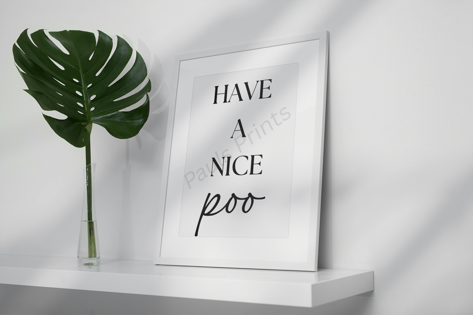 White framed humorous bathroom print with the text “Have a Nice Poo,” leaning on a shelf next to a tall green leaf in a glass vase.