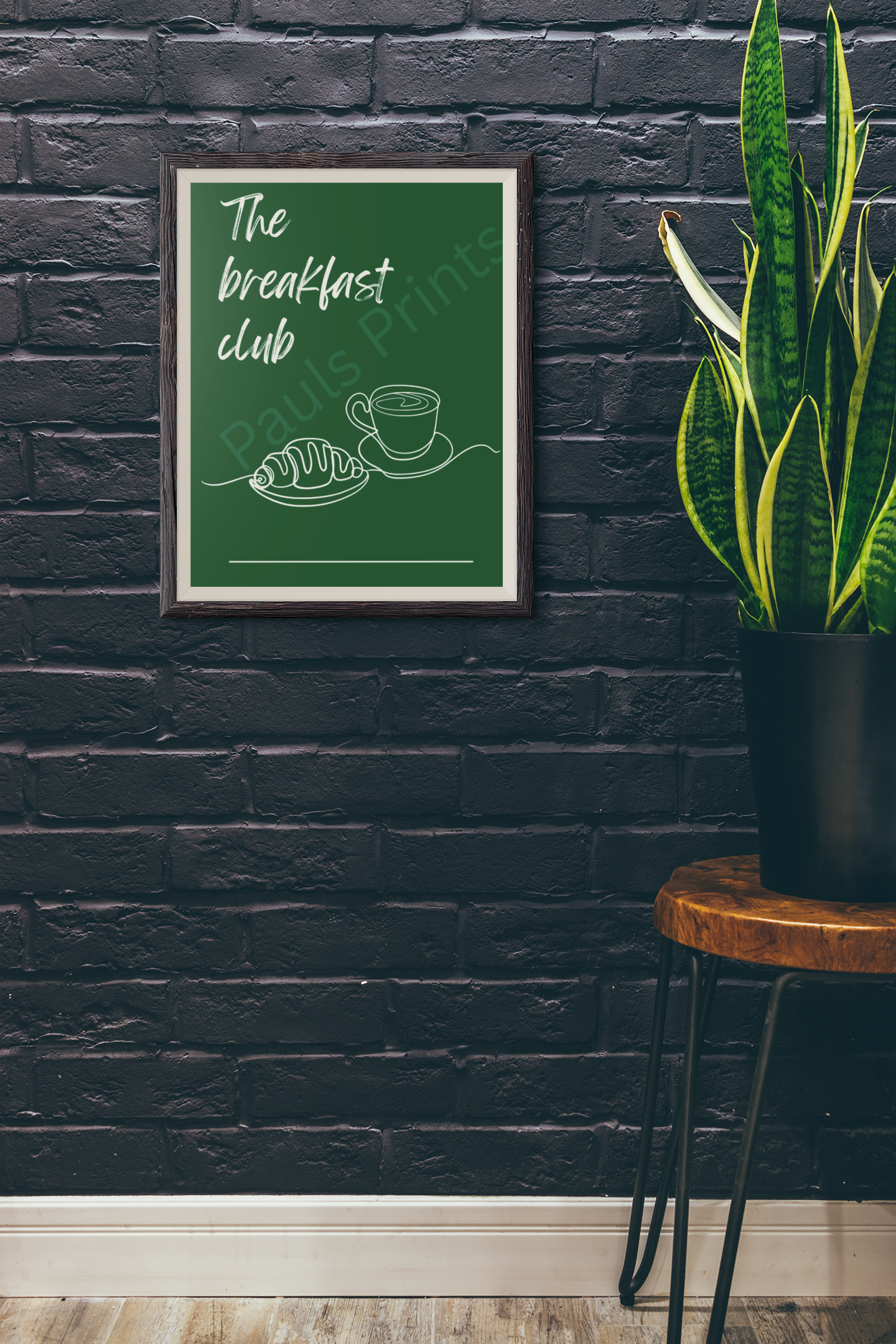 Framed "The Breakfast Club" print with croissant and coffee design on a dark brick wall, beside a snake plant in a rustic pot.
