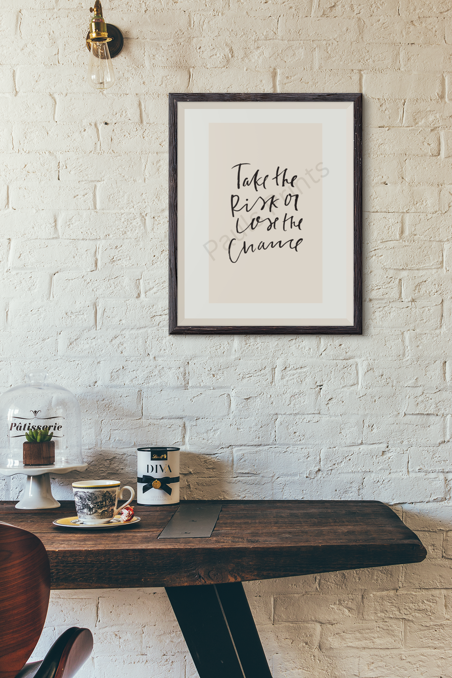 Rustic interior with white brick wall and framed print reading “Take the risk or lose the chance” in black handwritten script on beige background, above a wooden table with coffee cup.