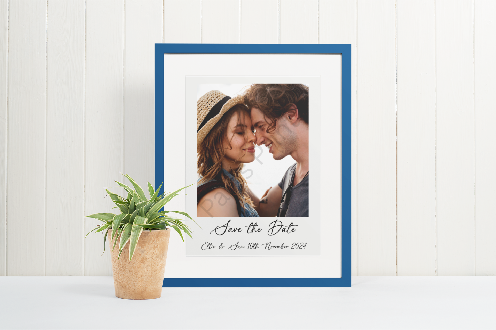 Blue-framed save the date print featuring a couple’s photo, styled against a white panel wall with a potted plant.