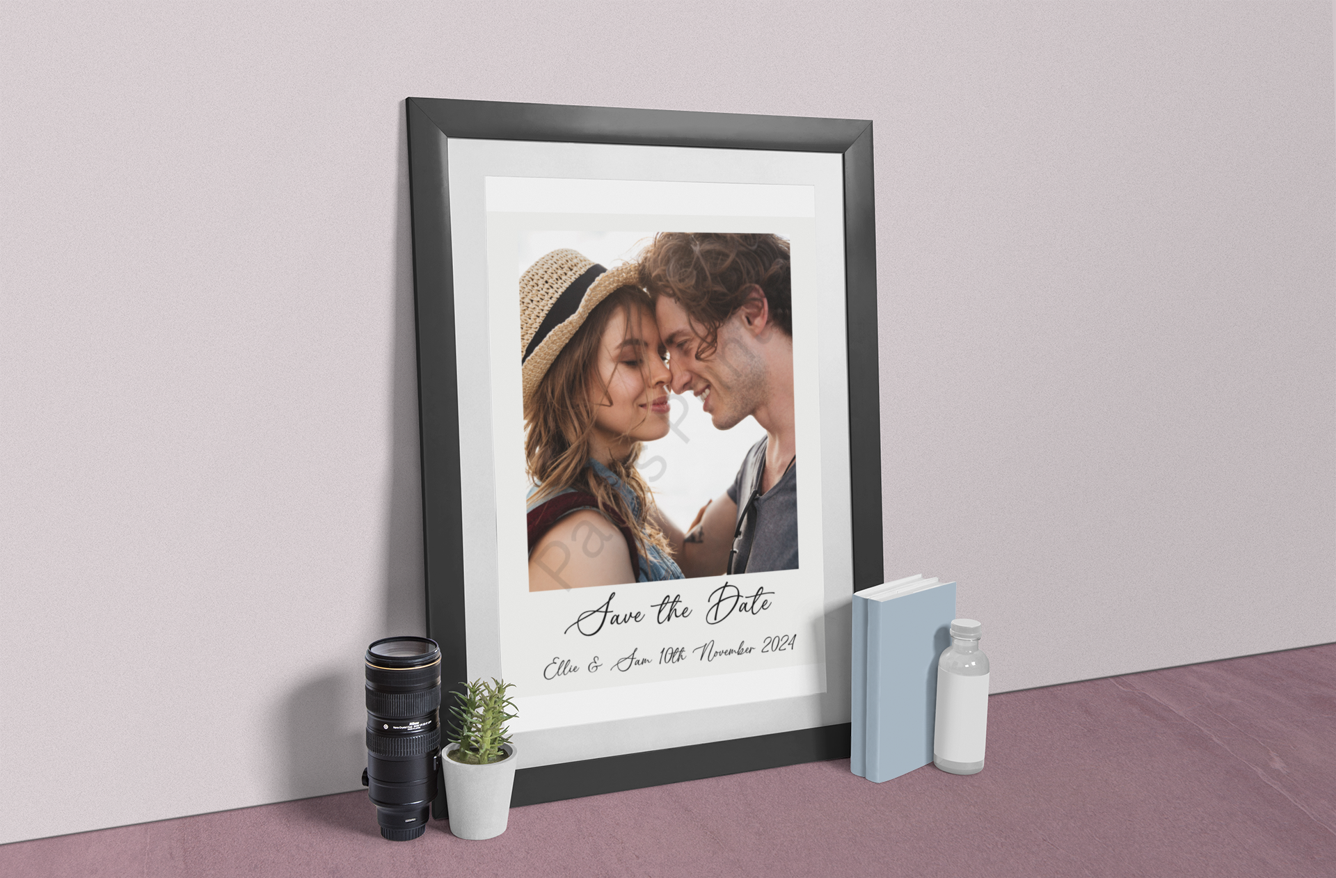 Black-framed save the date wedding print leaning against a wall on the floor, styled with camera lens, plant, and books.
