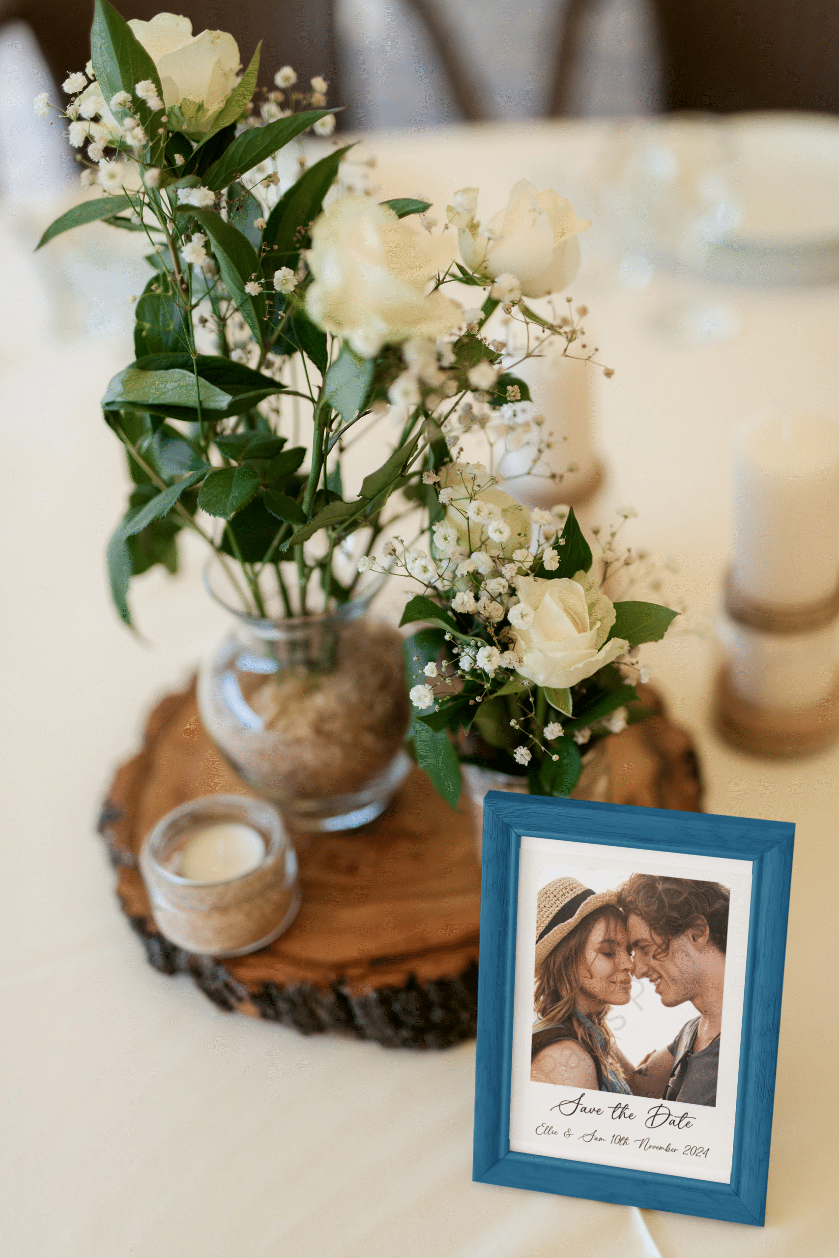 Blue-framed save the date wedding print on a table next to a floral centerpiece with white roses and candles.