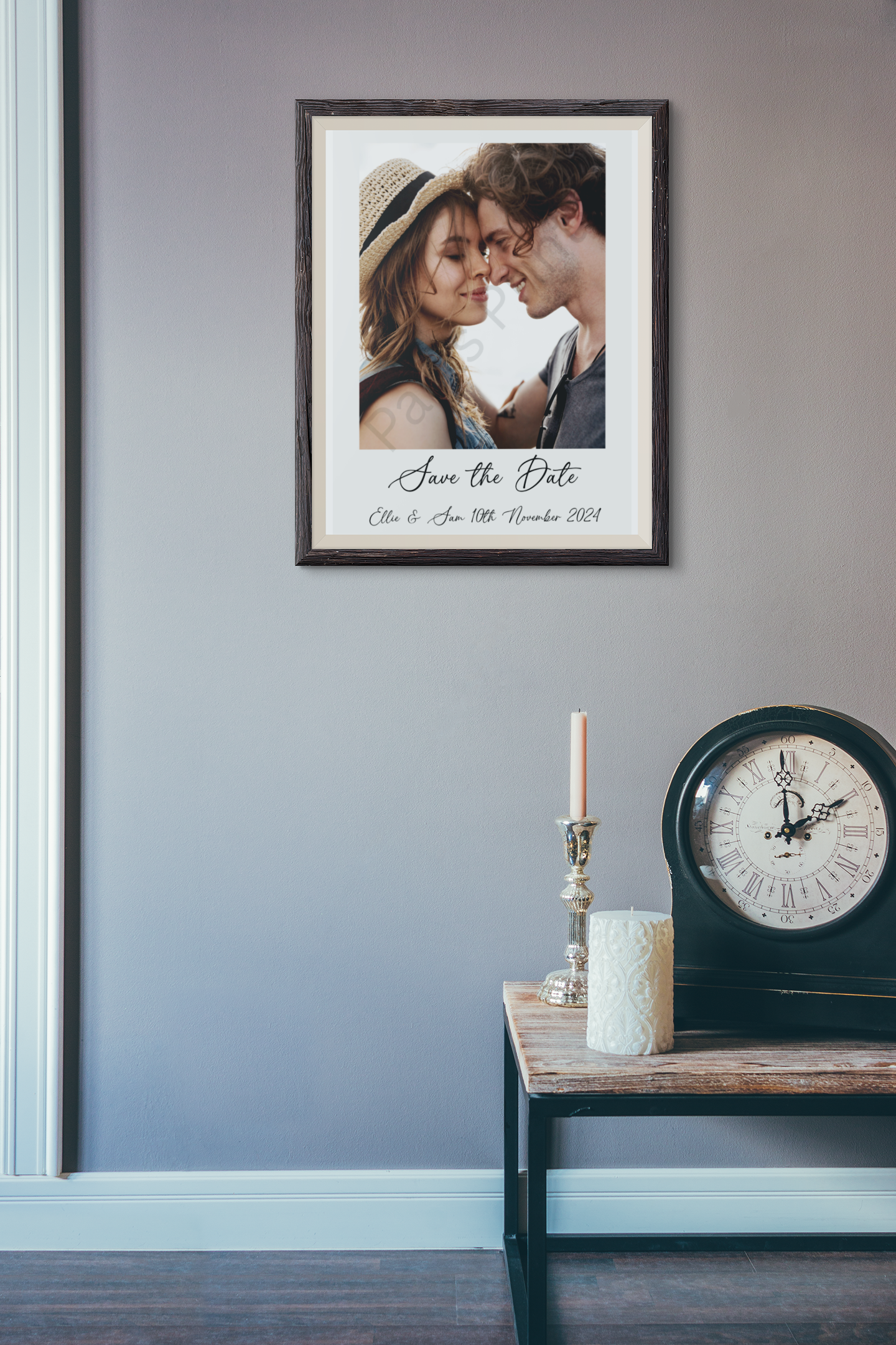 Save the date wedding print with a couple’s portrait in a rustic dark frame, hanging on a grey wall beside a table with a candle and clock.