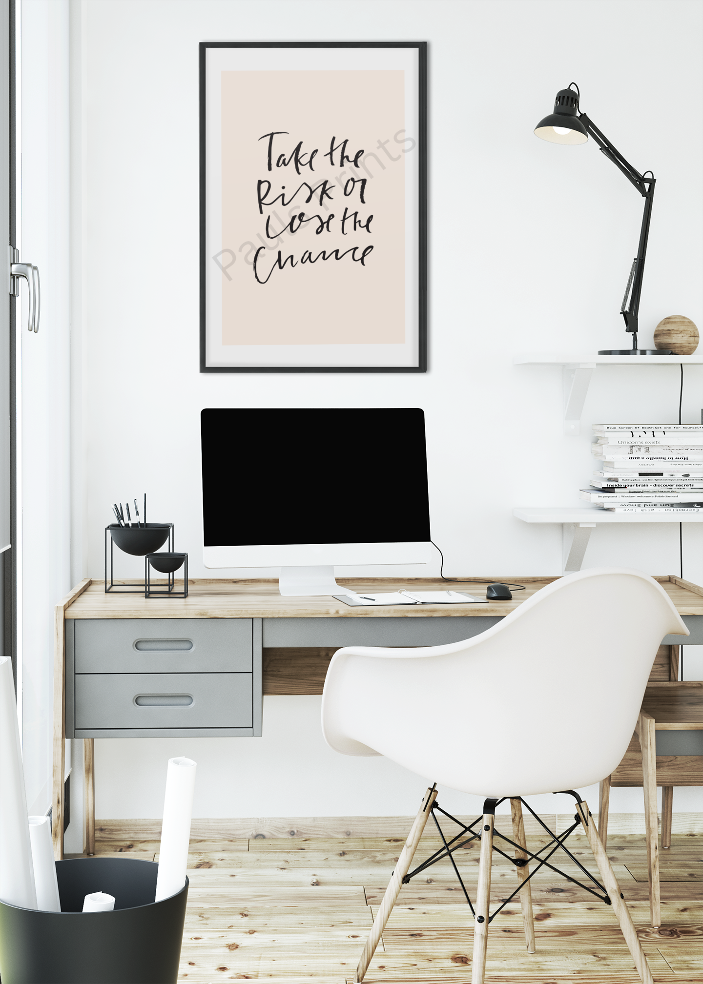 Modern office interior with framed motivational print reading “Take the risk or lose the chance” in black handwritten script on beige background, hung above a wooden desk with computer.