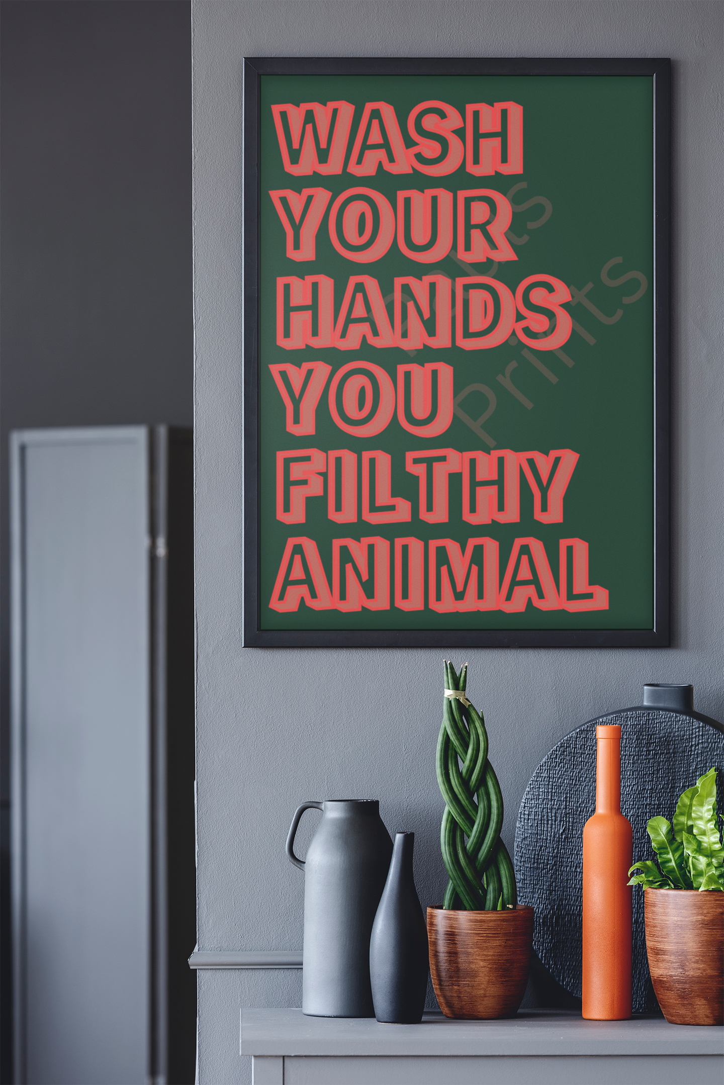 Playful typography print featuring red outlined bold letters on a green background reading “Wash Your Hands You Filthy Animal,” framed in light wood against a white textured wall.