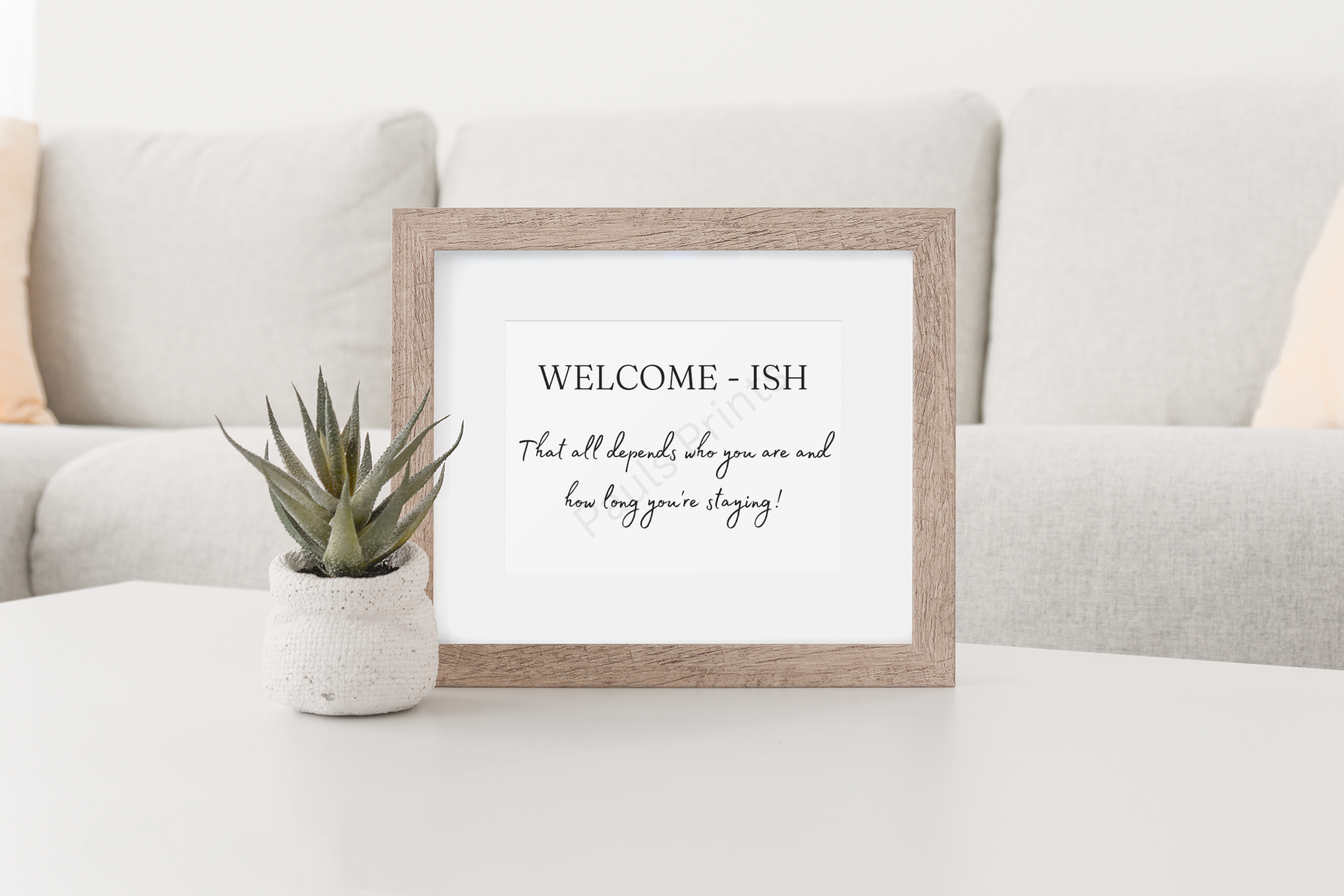 “Welcome-ish” humorous print in a wooden frame, placed on a white coffee table next to a small potted plant in a modern living room.
