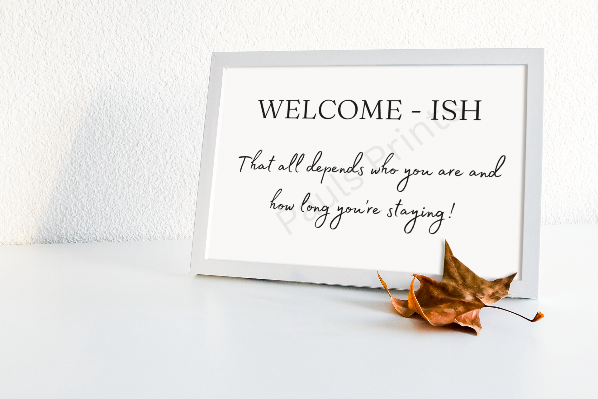 White framed print with the words "Welcome-ish – That all depends who you are and how long you’re staying!" placed on a white surface with a dried autumn leaf.
