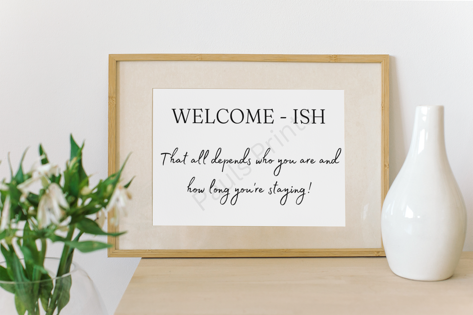 Playful “Welcome-ish” print in a wooden frame, leaning on a wall beside a white vase and green leafy plant.