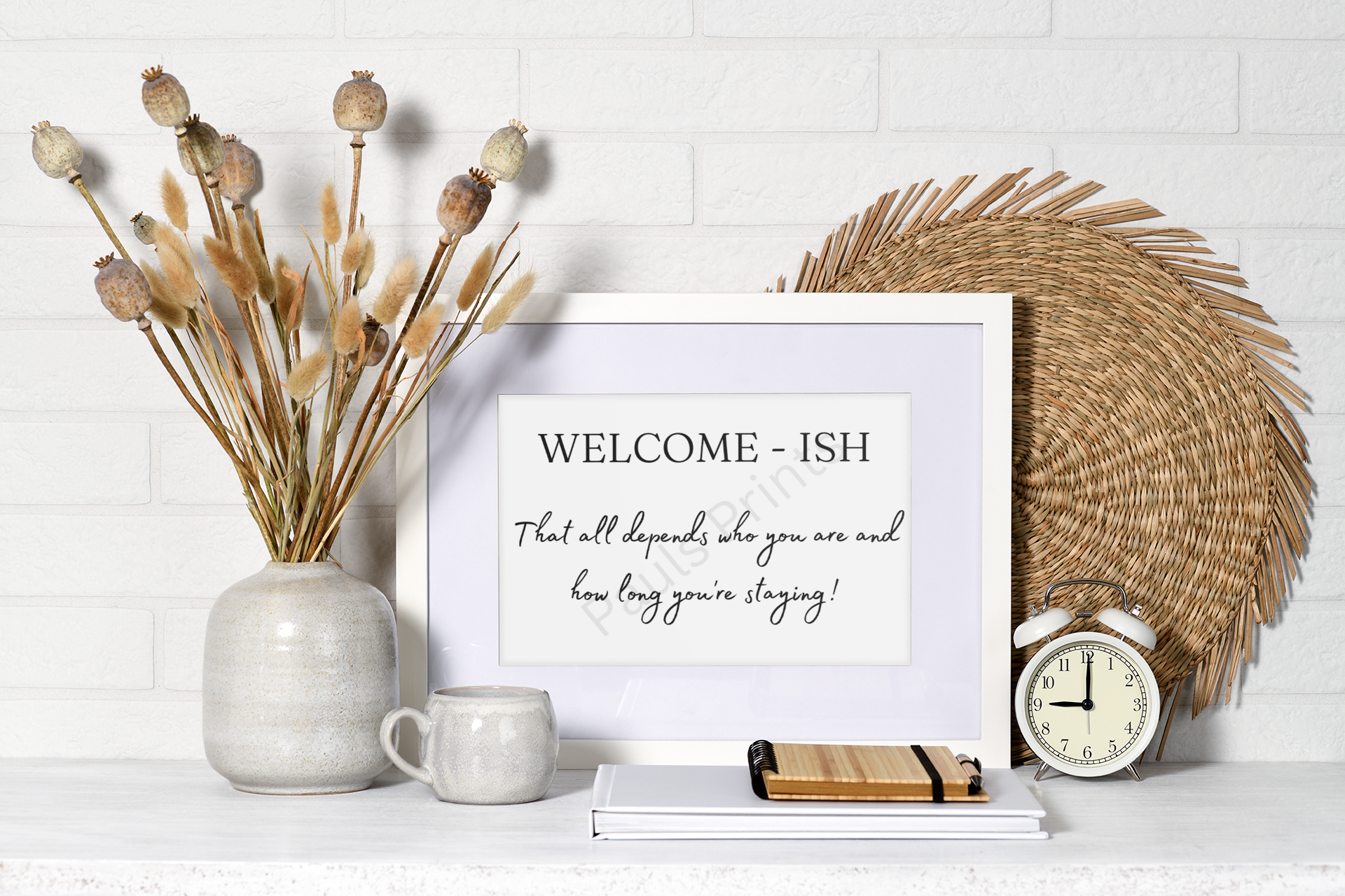 Framed “Welcome-ish” print with witty text, styled with neutral decor including dried plants, a woven placemat, a mug, and an alarm clock.