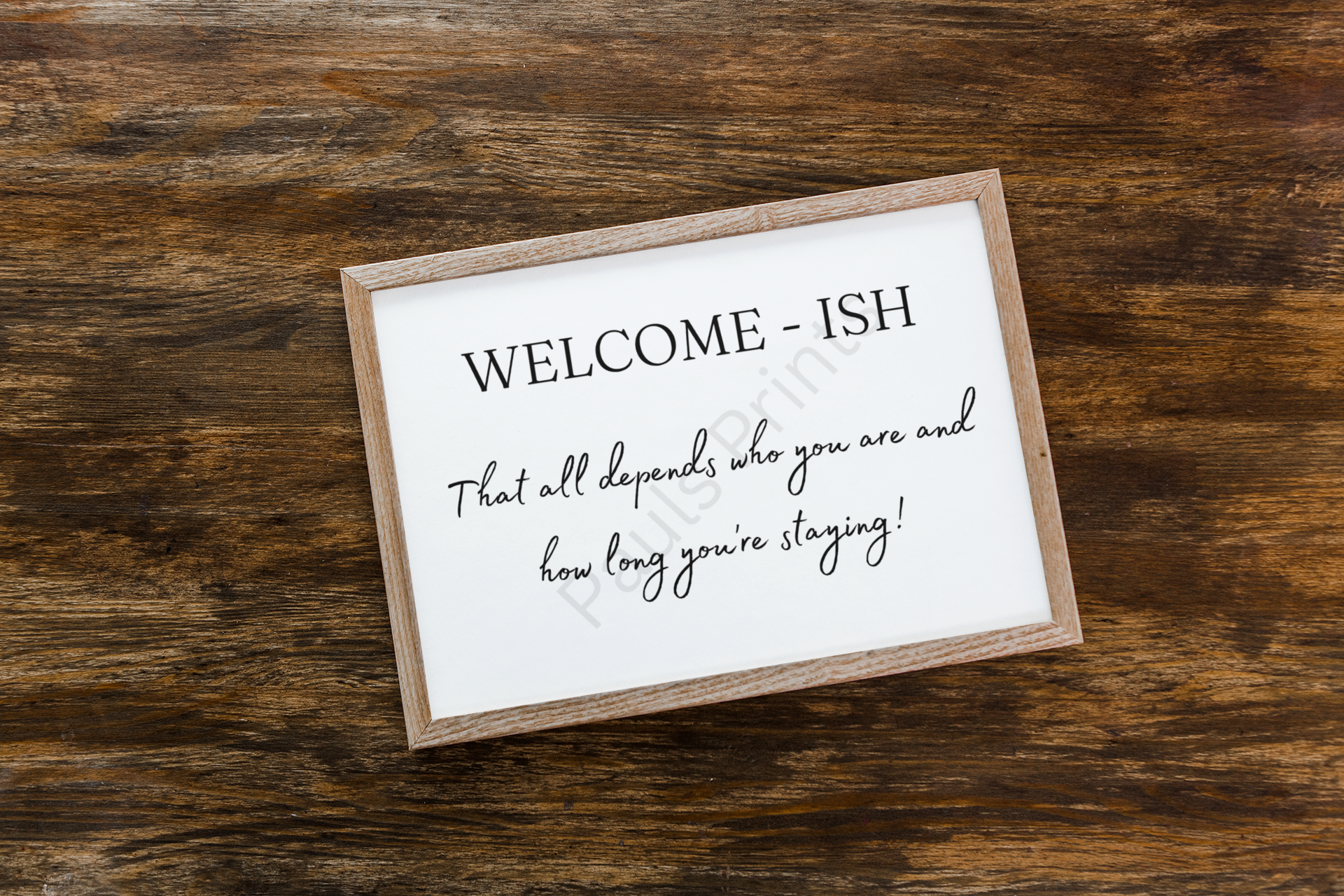 Wooden framed print with the text "Welcome-ish – That all depends who you are and how long you’re staying!" placed on a rustic wooden surface.